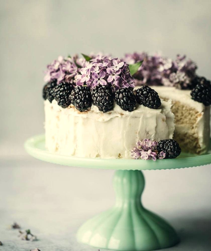 Vanilla celebration cake topped with blackberries and lilac flowers on a mint cake stand
