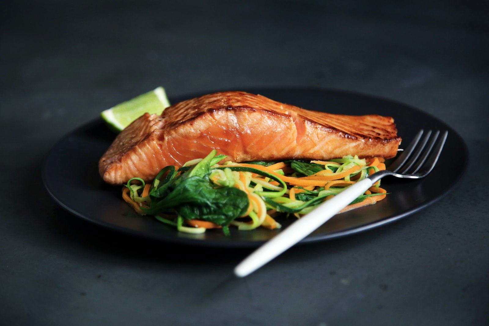 A seared fish fillet on a dark plate with ribbons of courgette and carrot, steam rising