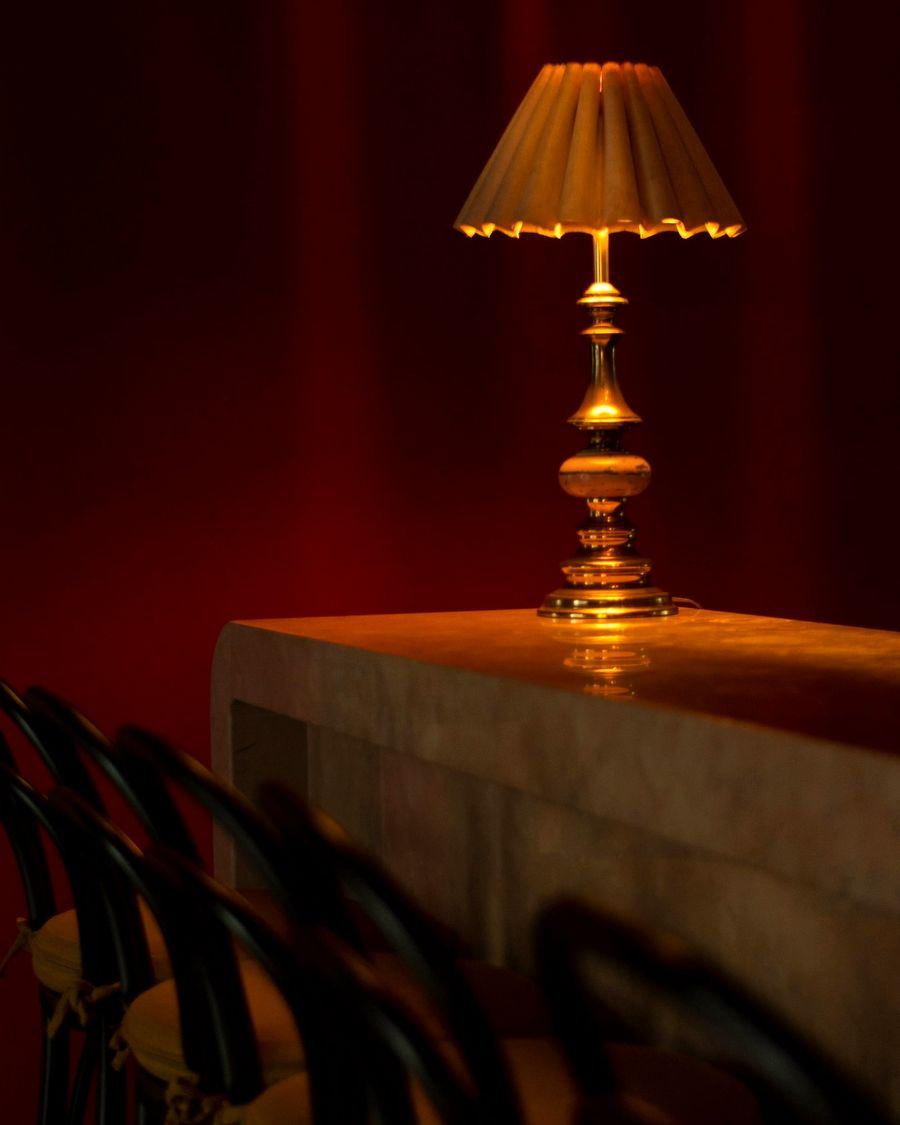 A turned brass table lamp with a pleated shade against an oxblood curtain
